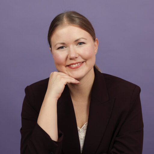 Iitu Leinonen nojaa korokkeeseen, katsoo kameraan ja hymyilee. Hänellä on päällään tumma bleiseri ja hiukset kiinni.