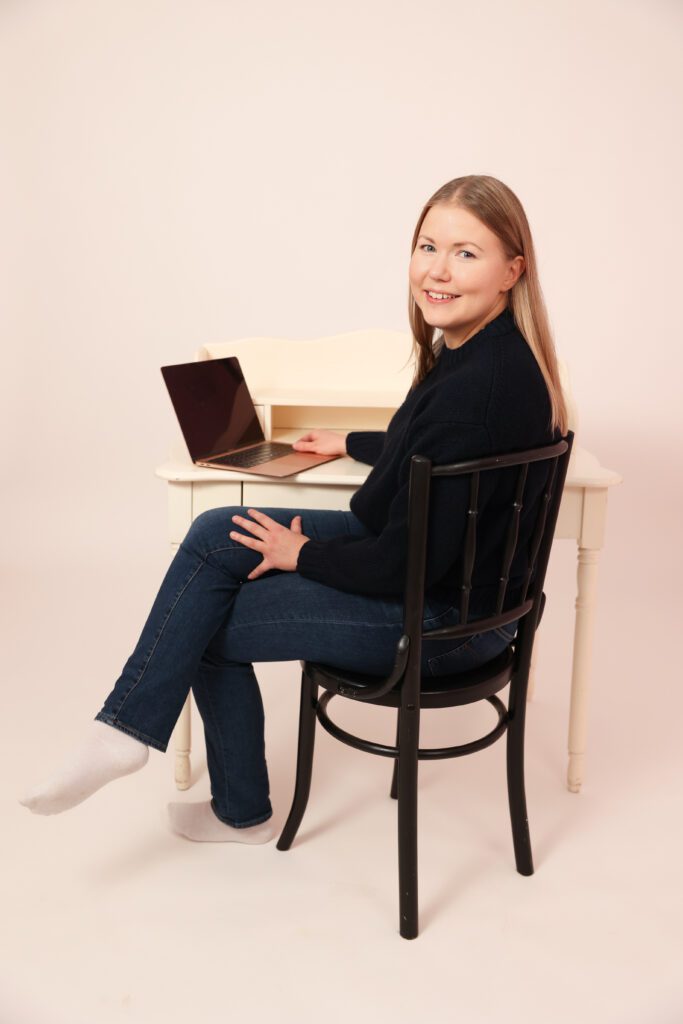 Iitu Leinonen istuu tuolilla ja katsoo kameraan hymyillen. Hänen edessään on pöytä, jolla on avoin tietokone.